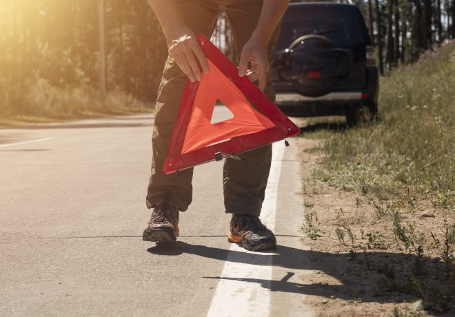 Male hands closeup putting red triangle caution sign on road side near broken car in summer.